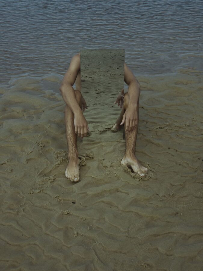 A person sits on wet sand with the ocean behind them, holding a rectangular mirror that hides their torso and head while their arms and legs frame the mirror’s reflection of the sand.