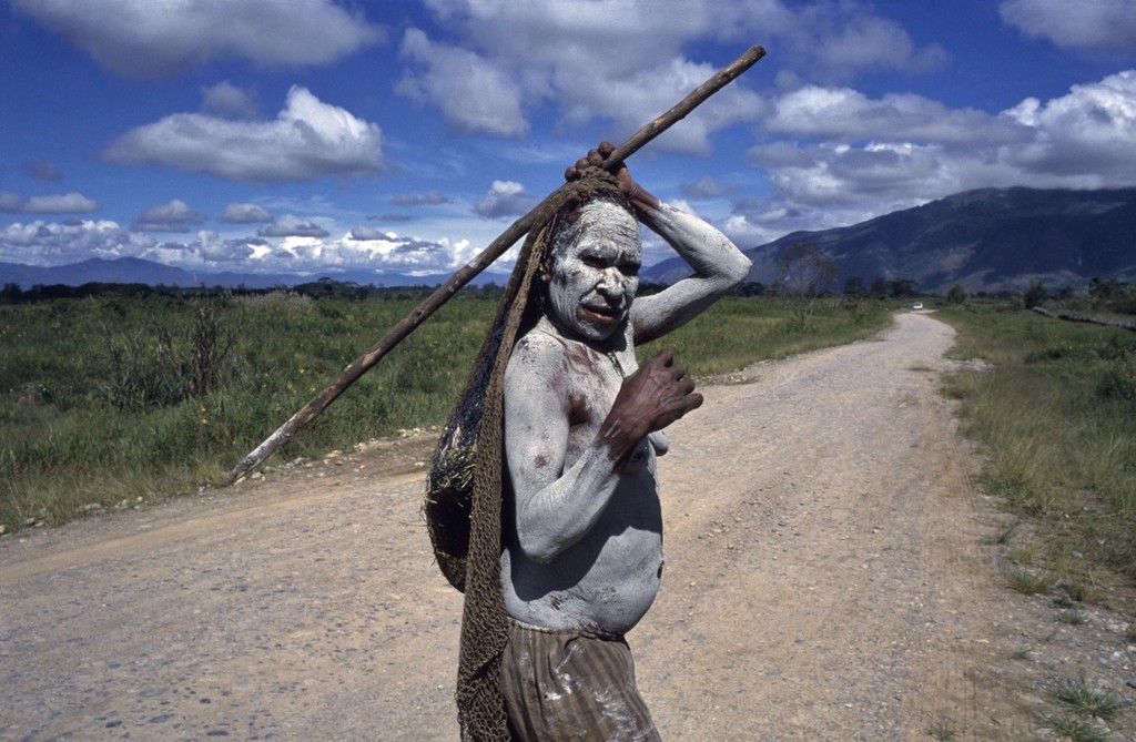 A person covered in white pigment walks along a dirt road in a wide valley with mountains rising in the distance, carrying a long wooden pole.