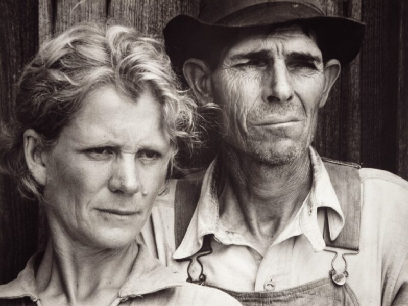 Black and white 1930s portrait of a rural couple standing close together against a wooden wall, their faces lined with strain as they look past the camera.