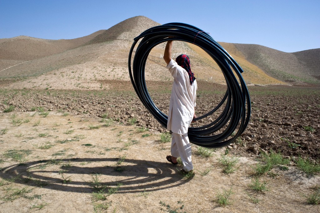 A person in loose-fitting clothing with a red head covering walking away from the camera into a desert landscape, carrying a large bundle of black plastic hose.