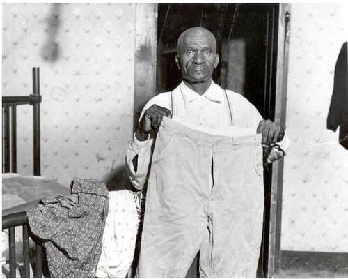 An older man stands in a modest room holding up a pair of trousers in front of him, looking directly at the camera. A bed with folded clothes is visible beside him.