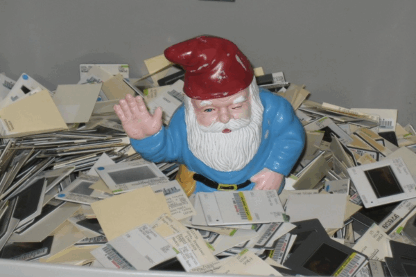 A garden gnome with a red hat and blue coat sits among piles of old photographic slides, waving with one hand.