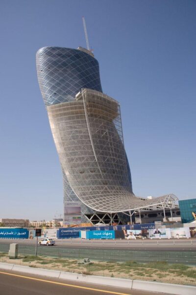 The Capital Gate building in Abu Dhabi, known for its distinctive leaning design. The glass-covered structure tilts at an impressive angle, showcasing its unique engineering. A clear blue sky forms the backdrop, with a road and construction signage visible in the foreground.
