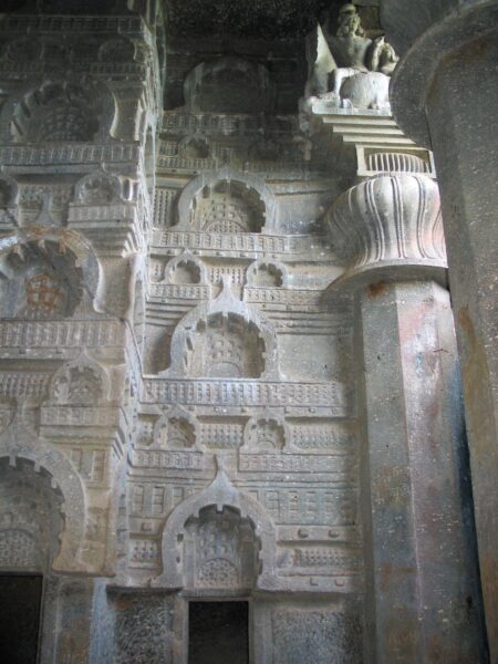 Intricate rock-cut carvings from the Bedsa Caves in Maharashtra, India, dating to the 1st century CE. The wall features a series of ornate chaitya windows, sculpted in repeating patterns, showcasing ancient architectural detailing. A decorative column with a lotus design and a partially visible sculpture of a seated figure are present in the upper corner, highlighting the artistic finesse of the era.