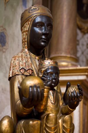 Close-up of a richly adorned statue of the Black Madonna and Child from the 11th century, featuring dark-toned faces and hands with golden robes and crowns. The Madonna holds a golden orb, symbolizing authority and divinity, while the Child also holds a spherical object. Intricate patterns and details on their garments highlight the craftsmanship, set against a softly lit ecclesiastical backdrop.