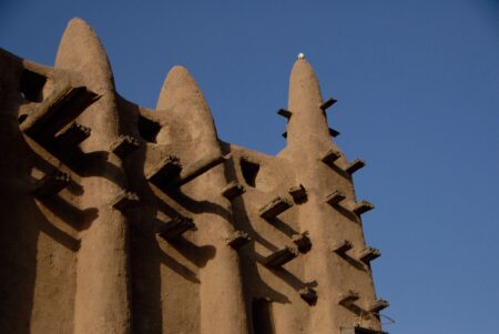 Close-up view of the Great Mosque of Djenné, showcasing its distinctive adobe architecture with large conical towers and protruding wooden beams known as torons. The earthy texture of the mud-brick structure contrasts against the deep blue sky, highlighting the unique design of this iconic Malian landmark, reconstructed in 1906.