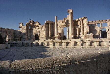 Exterior view of the ancient Roman theatre in Palmyra, Syria, seen from the south. The structure features columns and partially intact stone walls, showcasing classical Roman architectural elements. The ruins sit under a clear blue sky, with figures visible atop the remains, emphasizing the grandeur and historical significance of the site.