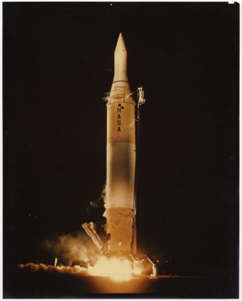 A nighttime photograph of the Juno II rocket launch by NASA. The rocket stands vertically, illuminated by flames and smoke at its base as it lifts off the ground. The NASA logo is visible on the rocket's body, and the dark sky forms the background.