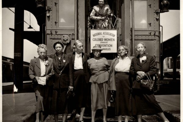 A black-and-white photograph of six African American women standing on a train platform in front of a railcar, with one woman leaning out of the train doorway. A sign on the train reads: “National Association of Colored Women Enroute Convention Los Angeles.” The women are dressed in formal attire, wearing suits, dresses, and hats, and carrying handbags. The image, taken between 1948-1952, captures a moment from the NACW’s journey to their convention.