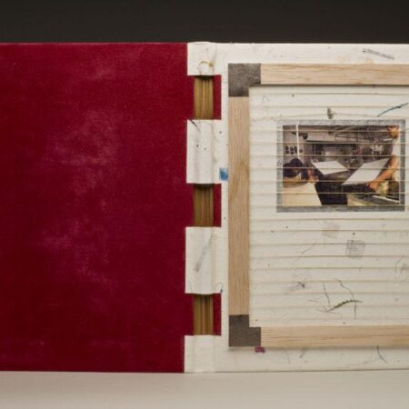A uniquely bound book with a white, speckled handmade paper cover and red suede interior. The cover features a recessed window containing a photograph of someone working in a print or bindery studio. The binding uses exposed wooden elements and visible stitching.