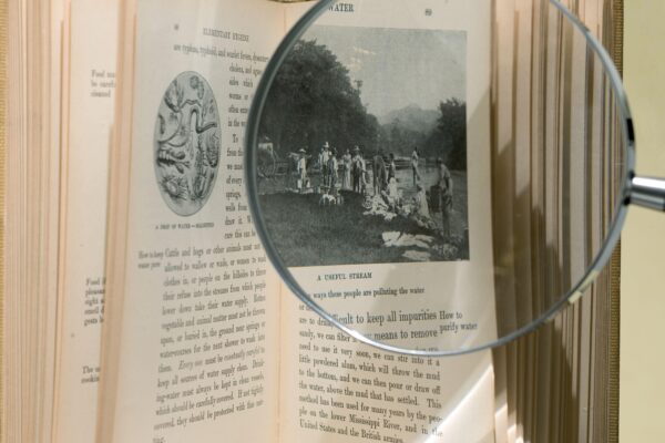 Image of an open book with a magnifying glass over an illustration titled "A useful stream", showing local residents and their livestock fetching water from a stream. Elementary hygiene for the tropics, p. 89.