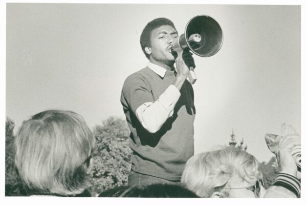 A black-and-white photograph of a man speaking passionately through a megaphone at an outdoor gathering. He wears a light shirt under a dark sweater and stands above a crowd, with trees visible in the background. Several people in the foreground, some with light-colored hair and glasses, listen closely and raise their hands.