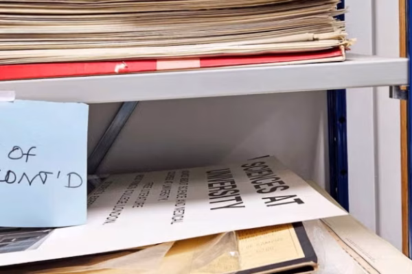 A cluttered metal shelf filled with stacks of old papers and folders. A blue sticky note taped to the shelf reads “SHELVES OF WOE, CONT’D” in black marker.