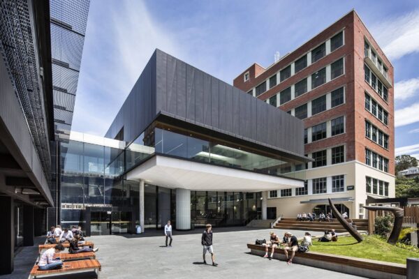 Modern campus courtyard at Victoria University of Wellington with students sitting and walking near the glass and brick Campus Hub and Library building.