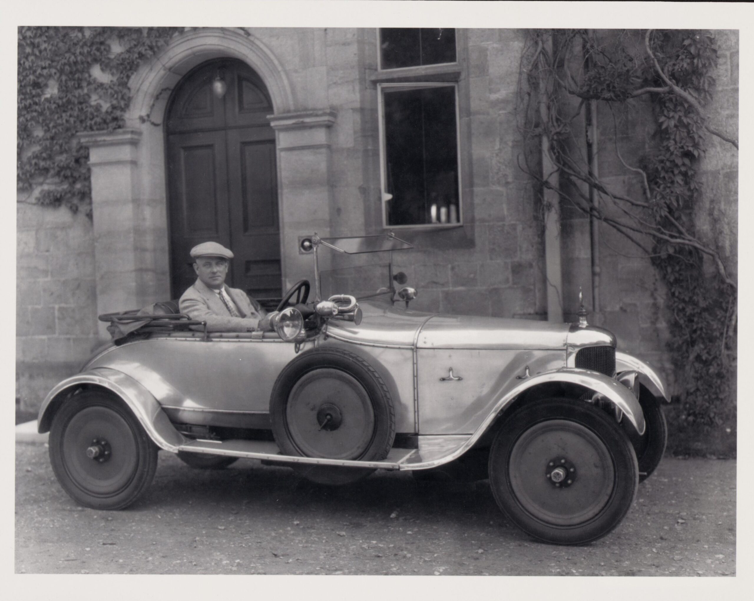 Black-and-white photo of a man in a flat cap seated in a small vintage open-top car, parked before a stone building with an arched doorway and ivy.