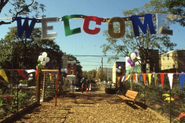A brightly decorated outdoor walkway leads into a community garden, with a large multicolored “WELCOME” banner hanging overhead. Balloons and colorful pennant flags line the fenced path, and people gather in the distance on a sunny day.