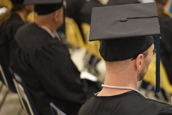 A room of people in graduation gowns and caps sitting with their backs to the camera, facing a speaker.