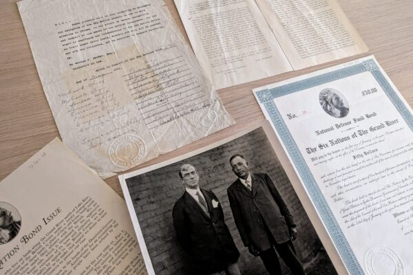 A flat lay of historical documents arranged on a wooden table, including typed and handwritten letters with embossed seals, printed booklets, a formal bond certificate with decorative blue border, and a black-and-white photograph of two men in early 20th-century suits standing against a brick wall. The papers appear aged and yellowed, suggesting archival materials.