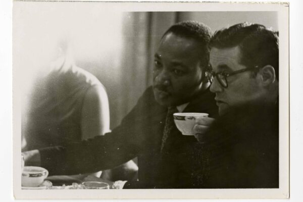 Black-and-white photograph of two men seated closely at a table, leaning forward and looking intently at something off-camera. The man in the center is Martin Luther King Jr., wearing a suit and tie, his expression focused. Beside him, another man wearing glasses holds a teacup near his mouth. A third figure is partially visible on the left, blurred by bright light. Cups and dishes sit on the table in the foreground, suggesting a quiet, candid moment during a meeting or conversation.
