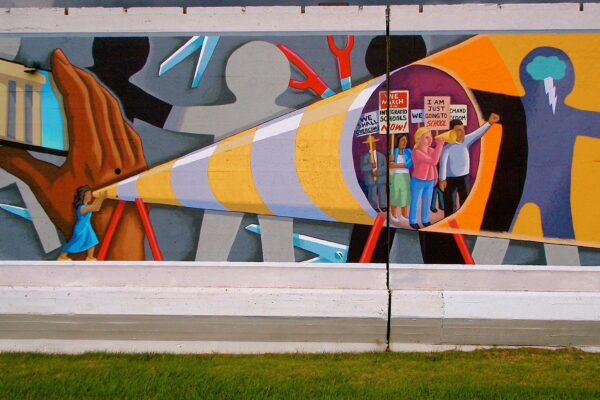 A large outdoor mural painted on a concrete wall depicts a sweeping civil rights and education justice narrative. At the center, a giant pencil rendered in yellow, white, and gray stripes extends diagonally across the composition, held at its base by a small figure in a blue dress. Inside the pencil's hollow tip, a group of diverse protesters carries signs reading "We March for Integrated Schools Now," "I Am Just Going to School," and "We Demand Freedom." To the left, figures march past a columned building , evoking the Lincoln Memorial, while a corn stalk and fire imagery appear below. To the right, large silhouetted figures in yellow and blue frame the scene, one with a lightning bolt emanating from their head. Scissors, tools, and fragmented shapes fill the background, suggesting barriers being cut through. The overall composition frames education and integration as acts of resistance and power. Sonnet 4.6