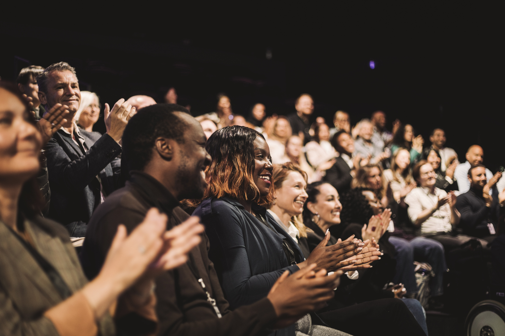 Audience members seated in a dim auditorium applauding during an event or presentation.