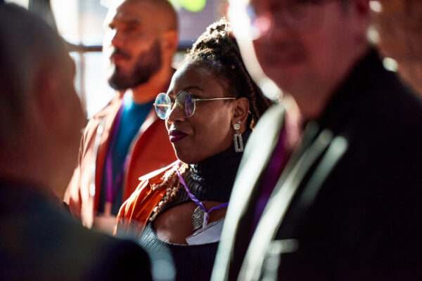 Attendees wearing conference badges engaged in conversation, with a woman in glasses in focus and others blurred in the foreground.