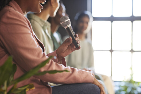 Person holding a microphone while speaking on a panel, with other participants seated nearby and a large window in the background.