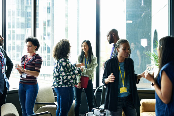 Group of people networking and talking in a bright room with large windows, some holding drinks and wearing event badges.