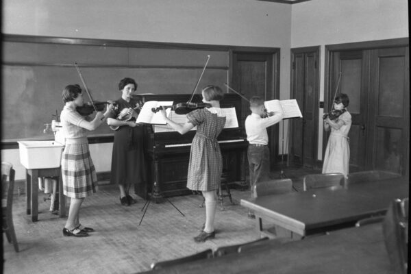 Black-and-white photograph of a music class, with an instructor standing beside a piano while four children play violins and read from music stands.