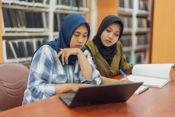 Two students study together at a library table, using a laptop and writing in a notebook.